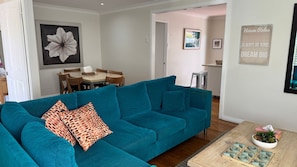 A teal sofa with giraffe-patterned pillows, a dining table with chairs, a kitchen area, and a wall with framed artwork and a 'House Rules' sign.