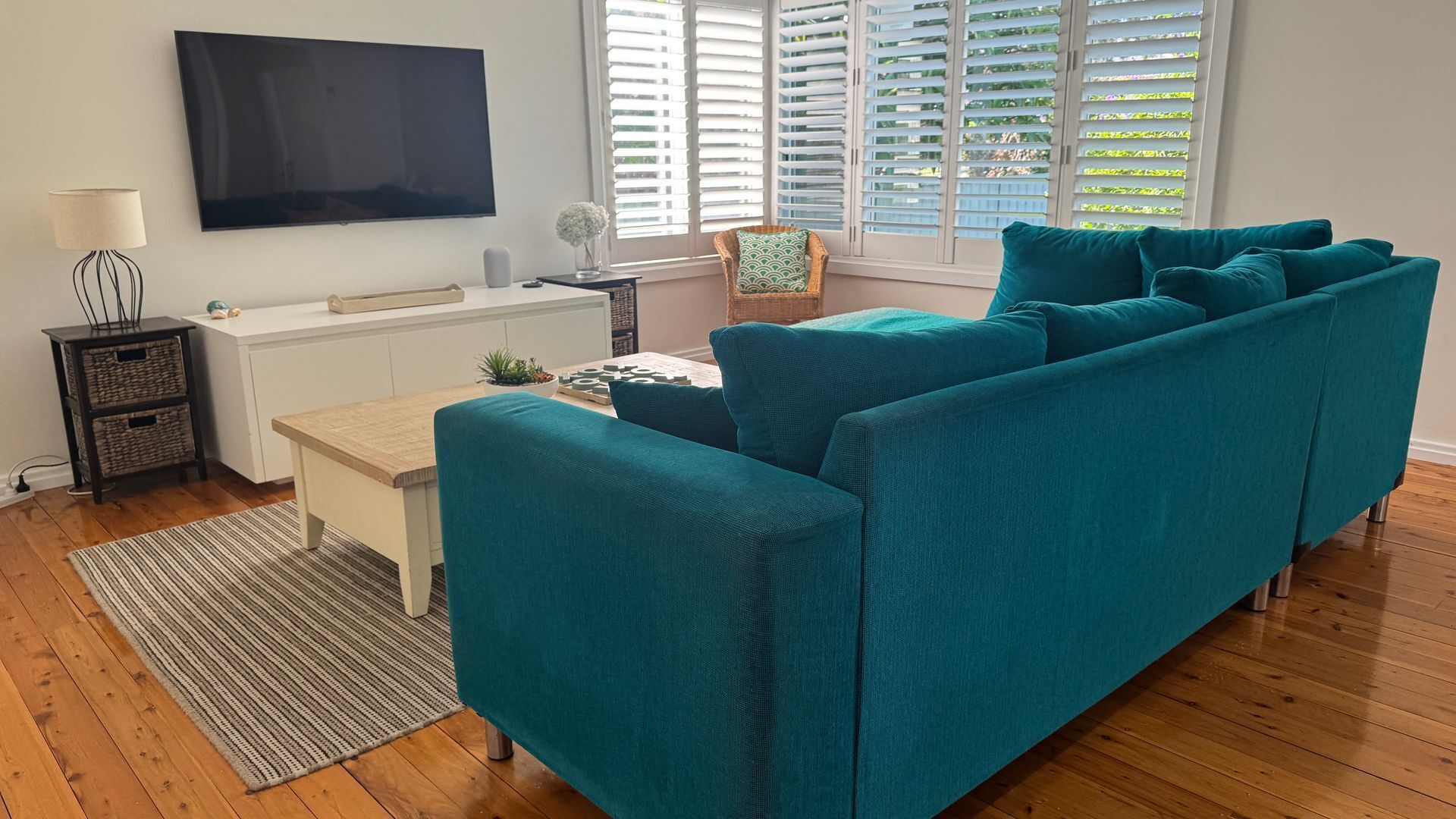A teal sofa in a living room with a flat-screen TV, a small table, and a window with blinds.