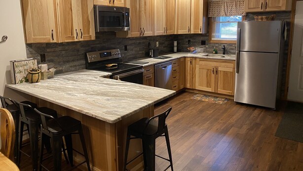 Una cocina moderna con armarios de madera, un refrigerador de acero inoxidable, un microondas, un horno y una encimera de mármol. Hay dos taburetes negros y un letrero de bienvenida.
