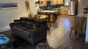 Amplia sala de estar con sofá, mesa de comedor y sillas. Se ve una cocina con muebles de madera y electrodomésticos al fondo.