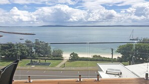Une vue depuis un balcon donnant sur une plage, avec des bateaux, un ciel dégagé et une côte lointaine.