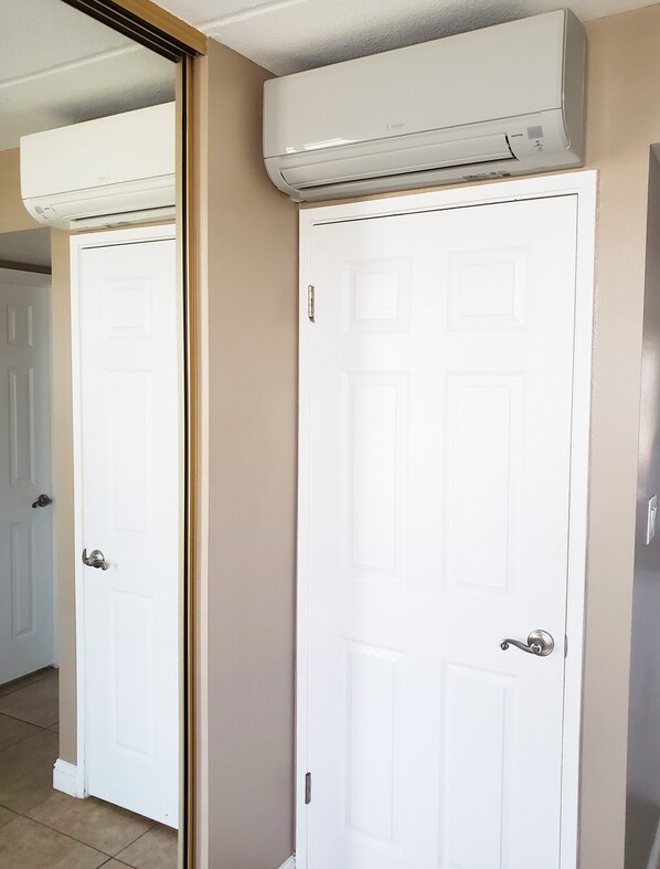 A white door with a silver handle, a white air conditioning unit mounted above it, and a mirror reflecting part of the room.