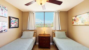 A room with two single beds, a ceiling fan, a nightstand with a lamp, and a window with curtains.