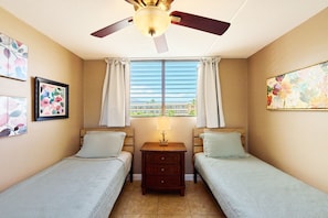 Una habitación con dos camas individuales, ventilador de techo, mesita de noche con lámpara y ventana con cortinas.