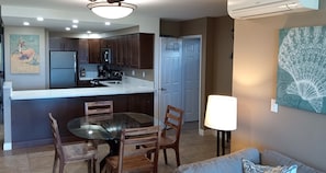 A modern kitchen with dark wooden cabinets, a stainless steel refrigerator, and a white countertop. There is a dining area with a glass table and wooden chairs. A lamp is on the floor, and a painting is on the wall.