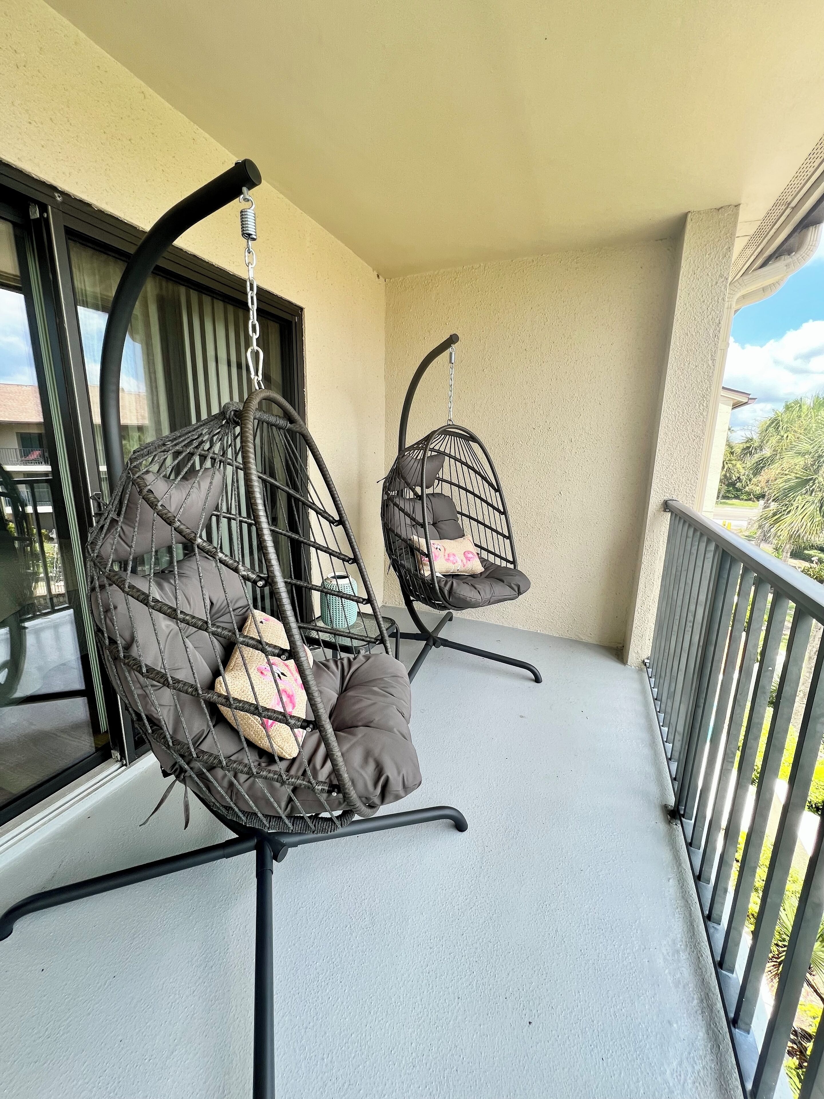 Two black wicker swing chairs with cushions on a balcony with a metal railing and a view of greenery.