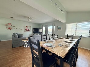 A dining area with a wooden table set for four, a gray sofa, a ceiling fan, and a TV on the wall. - The Flirty Flamingo 3br/2ba top floor condo in St. Augustine Beach (St Augustine Beach)