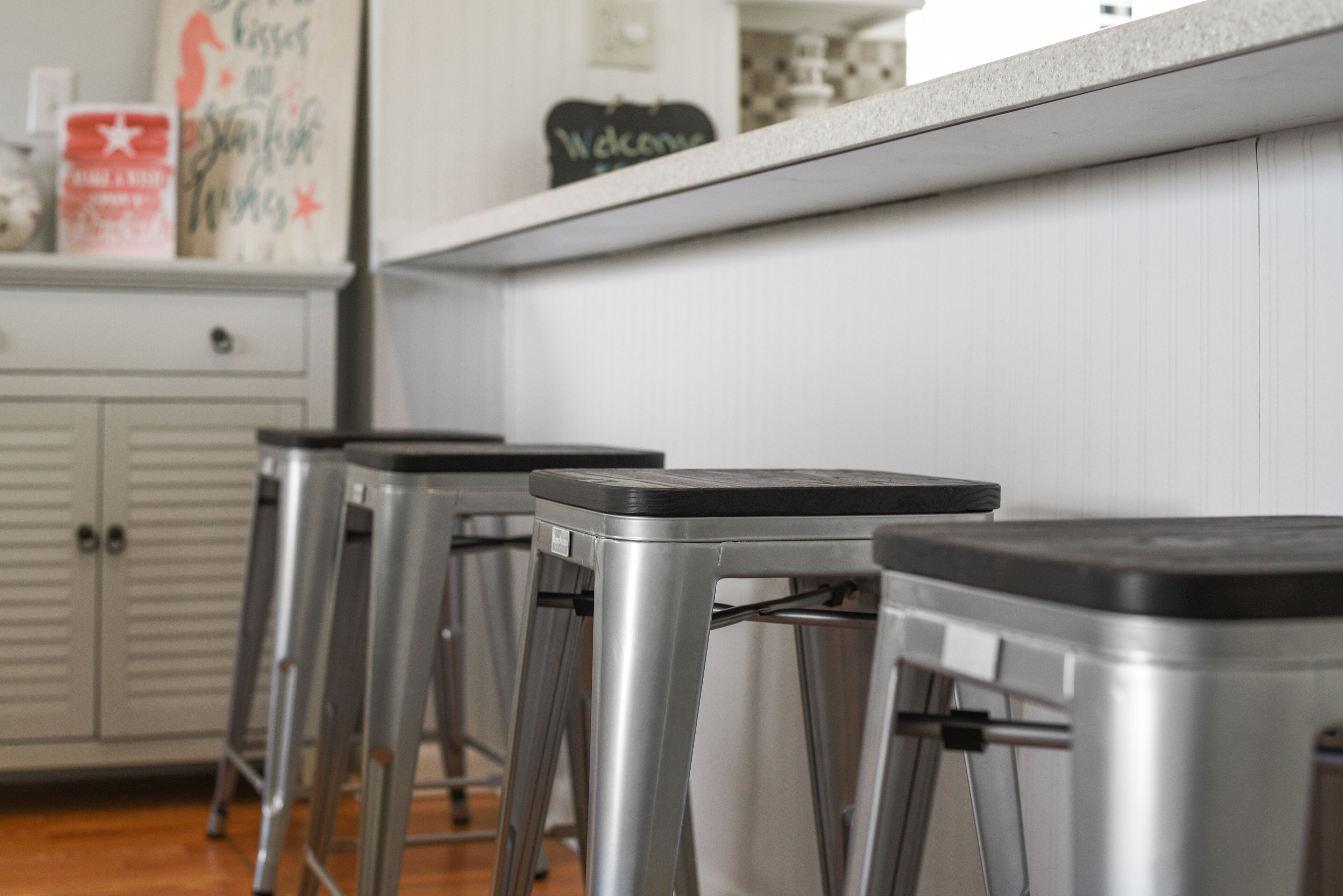 A kitchen with stainless steel bar stools, a white countertop, and a 'Welcome' sign.