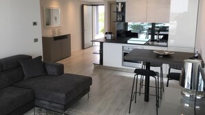 A modern living room with a dark grey sofa, a glass coffee table, and a kitchen area with a dining table and bar stools.