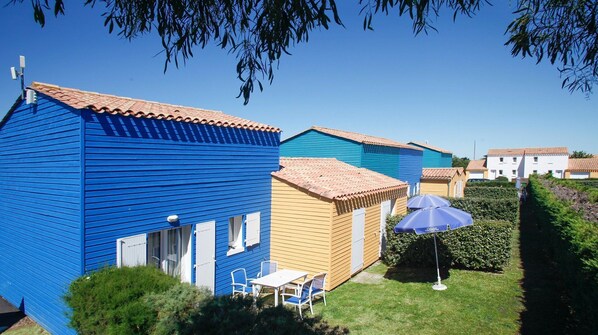 A row of colorful holiday cottages with tiled roofs, a small table with chairs, and a blue umbrella.