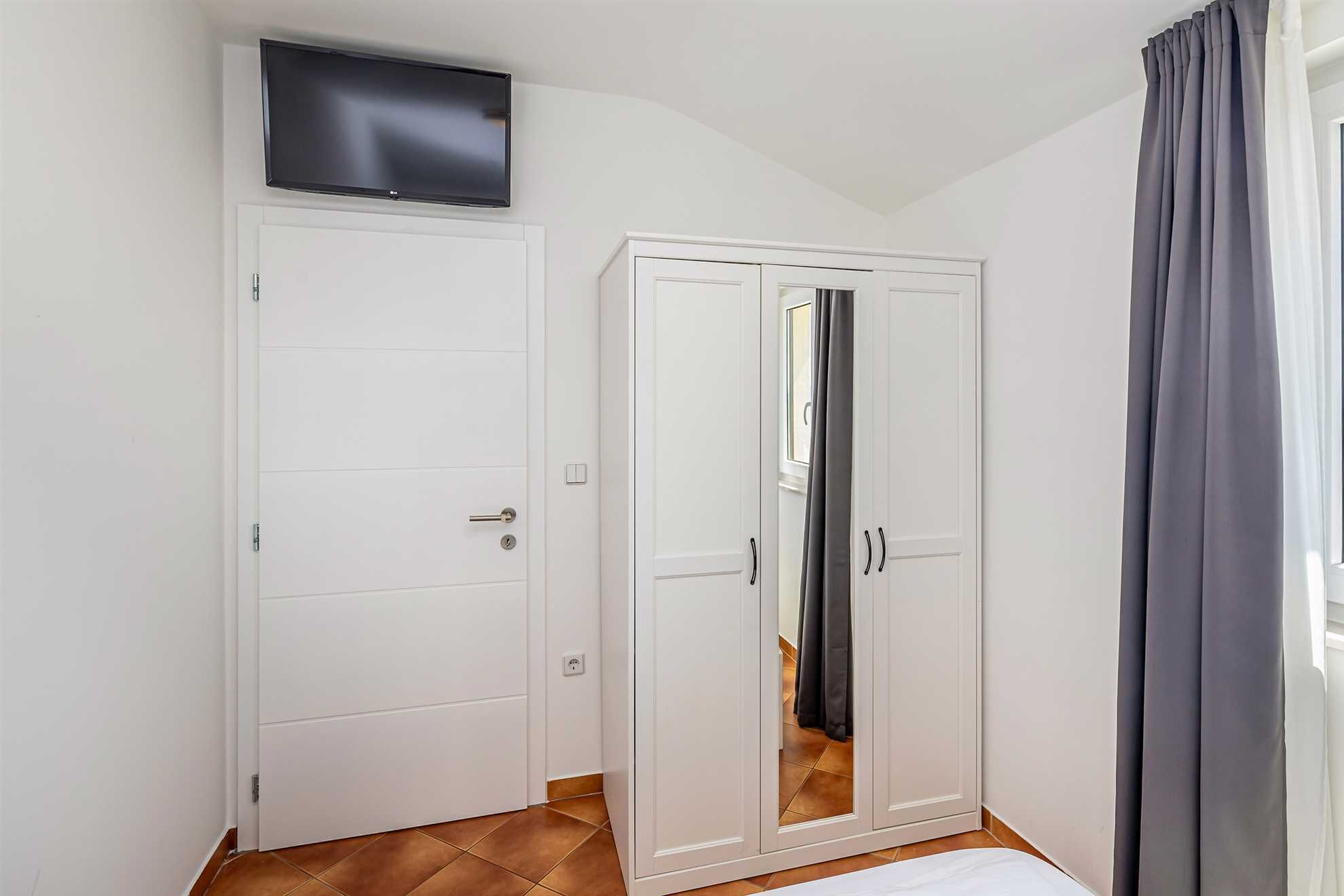 Een kamer met een witte kast, een wandtelevisie en een raam met grijze gordijnen.