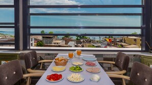Une table dressée avec des aliments pour le petit-déjeuner, et une vue sur une ville côtière à travers de grandes fenêtres.