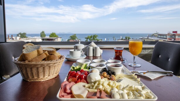 Une table de petit-déjeuner garnie de divers aliments et un verre de jus d’orange, avec une vue panoramique sur la mer et les bâtiments.