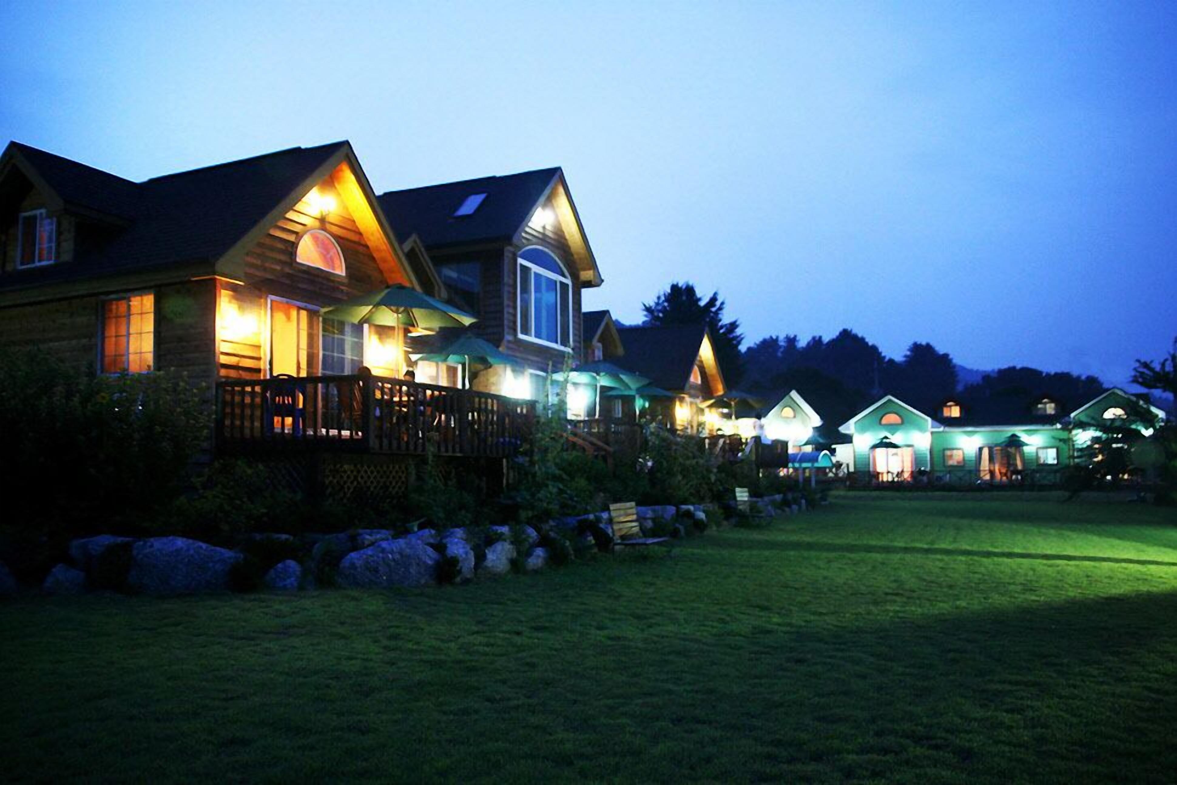 A row of log cabin-style houses with wooden decks and large windows, surrounded by a well-maintained lawn and a stone wall.