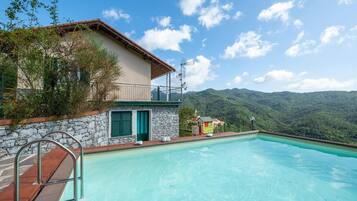 Vista della piscina, con un edificio, un muro in pietra, un balcone e montagne sullo sfondo.