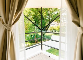 Ein Zimmer mit einem großen Fenster, das einen geometrischen Metallrahmen, zur Oberseite gebundene Vorhänge und Blick auf einen grünen Garten und Bäume hat.