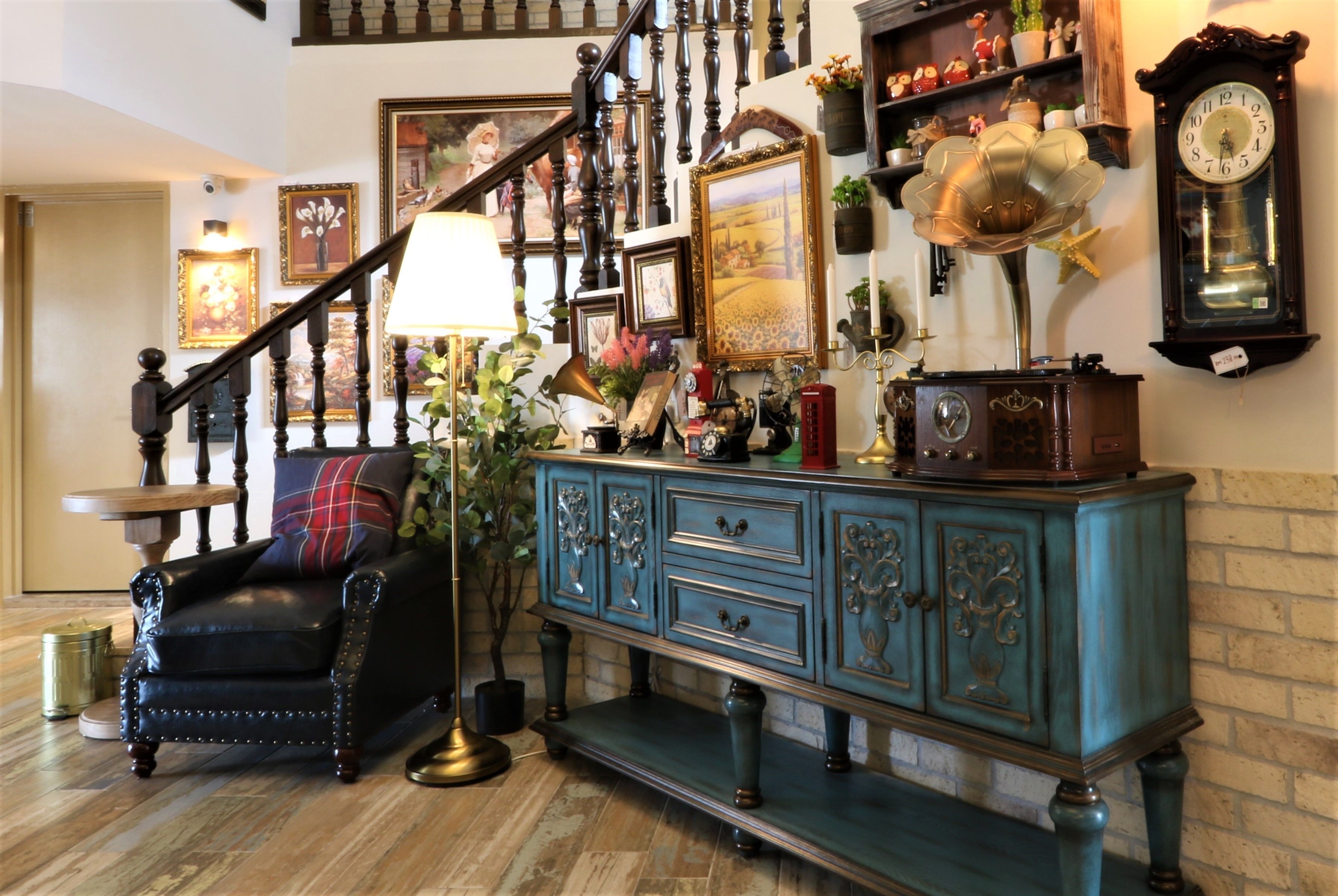 A staircase with ornate railings, a black leather armchair with a plaid cushion, a teal dresser with decorative carvings, and a wall clock.