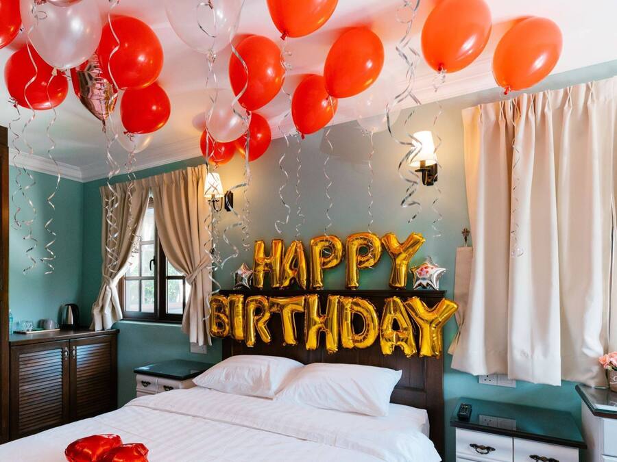 Ett sovrum med en säng, ett nattduksbord, en byrå och ett fönster med gardiner. Det finns ballonger och en skylt med texten ”Grattis på födelsedagen” på väggen.
