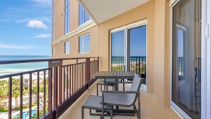 A balcony with a table and chairs, overlooking a beach and pool area.