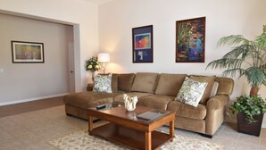 A living room with a brown sofa, a glass coffee table, and a patterned rug. There are two framed artworks on the wall and a potted plant.