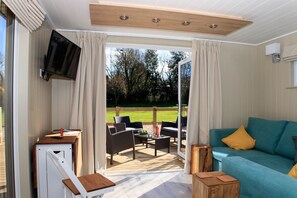 A room with a blue sofa, a television mounted on the wall, and a sliding glass door leading to an outdoor patio area. - Luxury Contempoary Lodge Nr Bath & Bristol (Bishop Sutton)
