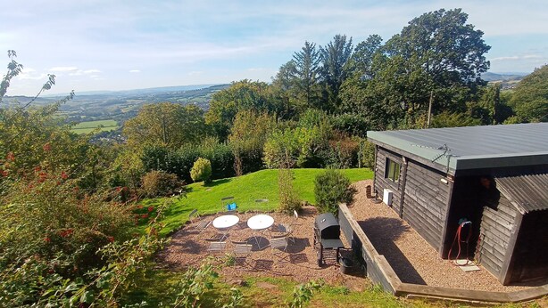 A wooden cabin with a gravel patio, surrounded by lush greenery and a scenic view of rolling hills.