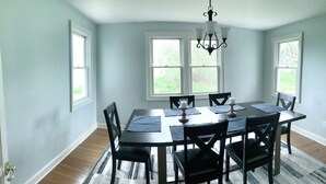 Uma sala de jantar com uma mesa de madeira escura, seis cadeiras, um candelabro e grandes janelas com vista para o verde.