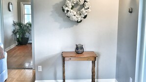 Uma sala de estar com piso de madeira, parede cinza claro, grinalda floral branca e uma pequena mesa de madeira com um objeto decorativo em cima.