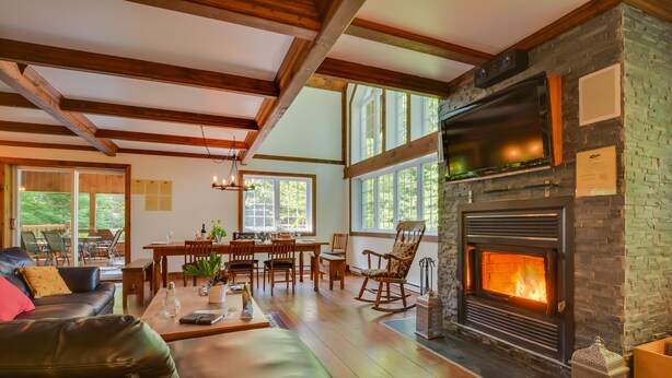 A cozy living room with a fireplace, wooden beams, a dining area, and a view of the outdoors. - Chalet Le Supérieur/Tremblant- Lac-Supérieur/Spa++ (Lac-Supérieur)