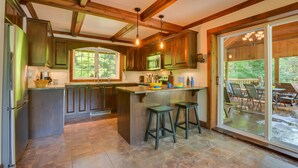 A kitchen with wooden beams, green cabinets, a central island with stools, and a view to an outdoor patio. - Chalet Le Supérieur/Tremblant- Lac-Supérieur/Spa++ (Lac-Supérieur)