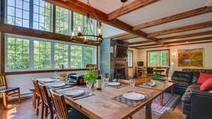A spacious dining area with a large wooden table set for six, surrounded by wooden chairs. A stone fireplace and a television are visible in the background. - Chalet Le Supérieur/Tremblant- Lac-Supérieur/Spa++ (Lac-Supérieur)