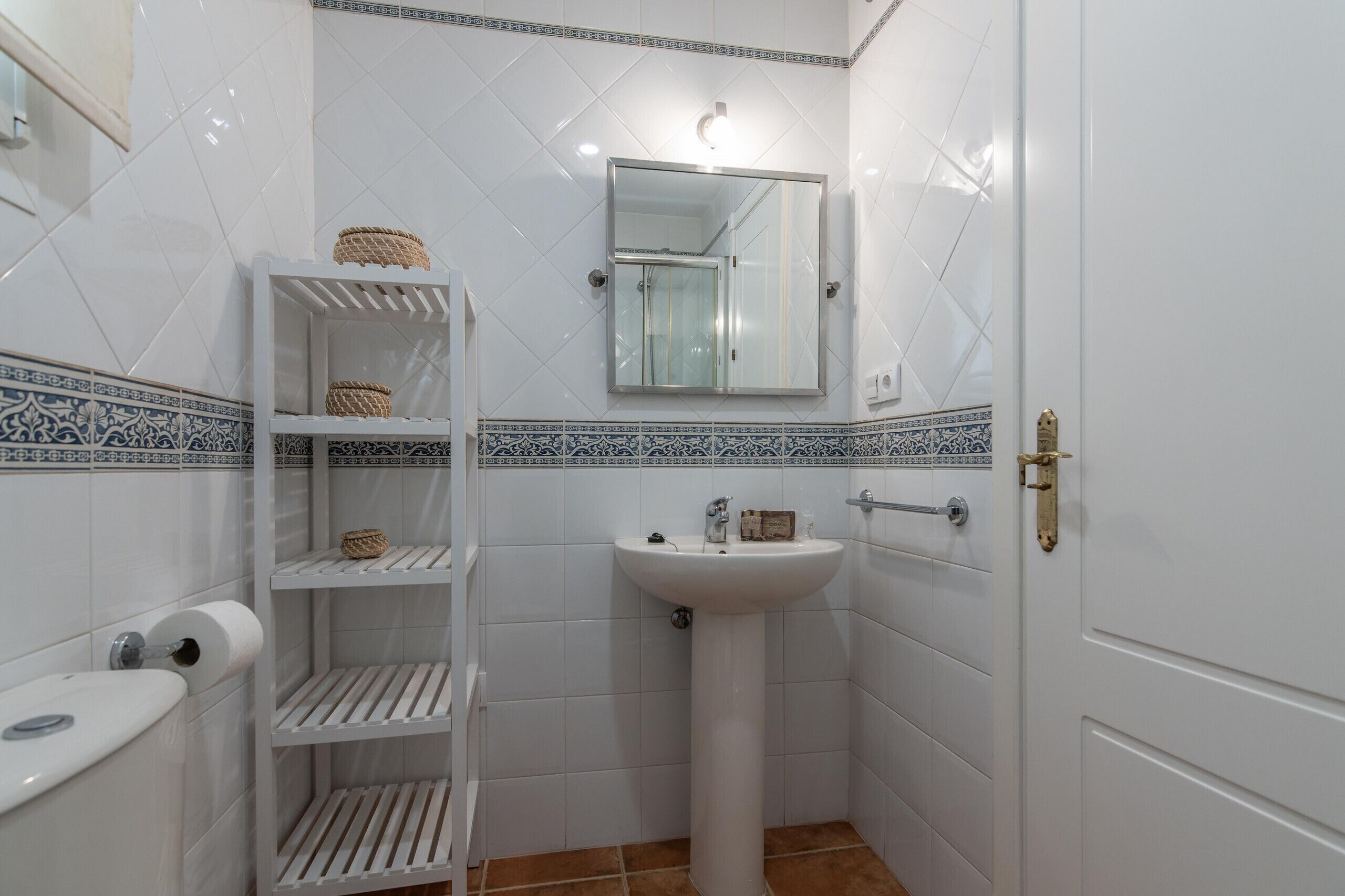 A bathroom with white tiled walls, a white sink, a mirror, and a shelf with baskets.