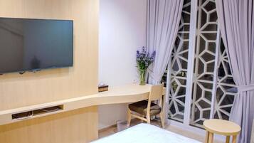 A modern hotel room with a flat-screen TV mounted on the wall, a wooden desk with a chair, a small round table, and a bed with white bedding.
