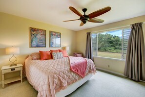 Une chambre avec un grand lit, un ventilateur de plafond, deux tableaux accrochés au mur, une fenêtre avec des rideaux et une table de chevet avec une lampe.