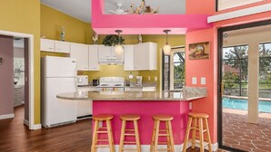 Een keuken met een roze accentmuur, witte kasten, een centraal eiland met krukken, en een zwembad zichtbaar door een glazen deur.