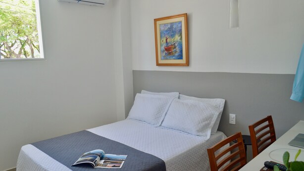 Una cama bien hecha con una revista, un cuadro en la pared y una ventana con vistas a árboles.