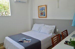 A neatly made bed with a magazine, a painting on the wall, and a window with a view of trees. - Pousada Praia Pajuçara (Maceió)