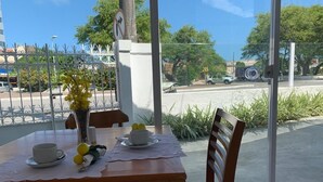 Un área de comedor al aire libre con una mesa para dos, un florero con flores amarillas y vista a una calle con coches y árboles.