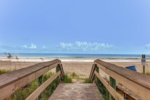 Ein hölzerner Steg führt zu einem Strand mit freiem Blick auf das Meer und einem Wegweiser in der Ferne.