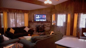 A cozy living room with a fireplace, a television, and a kitchen area. - Spacious, bright, cozy, single floor home nestled in pine trees (Groveland)