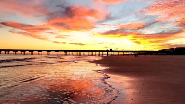 Ein Strand mit einem Steg, der sich ins Wasser erstreckt, sanfte Wellen, die an das Ufer schlagen, und ein Sonnenuntergang im Hintergrund.