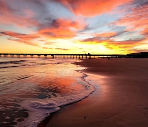 A beach with a pier extending into the water, waves gently lapping at the shore, and a sunset in the background.