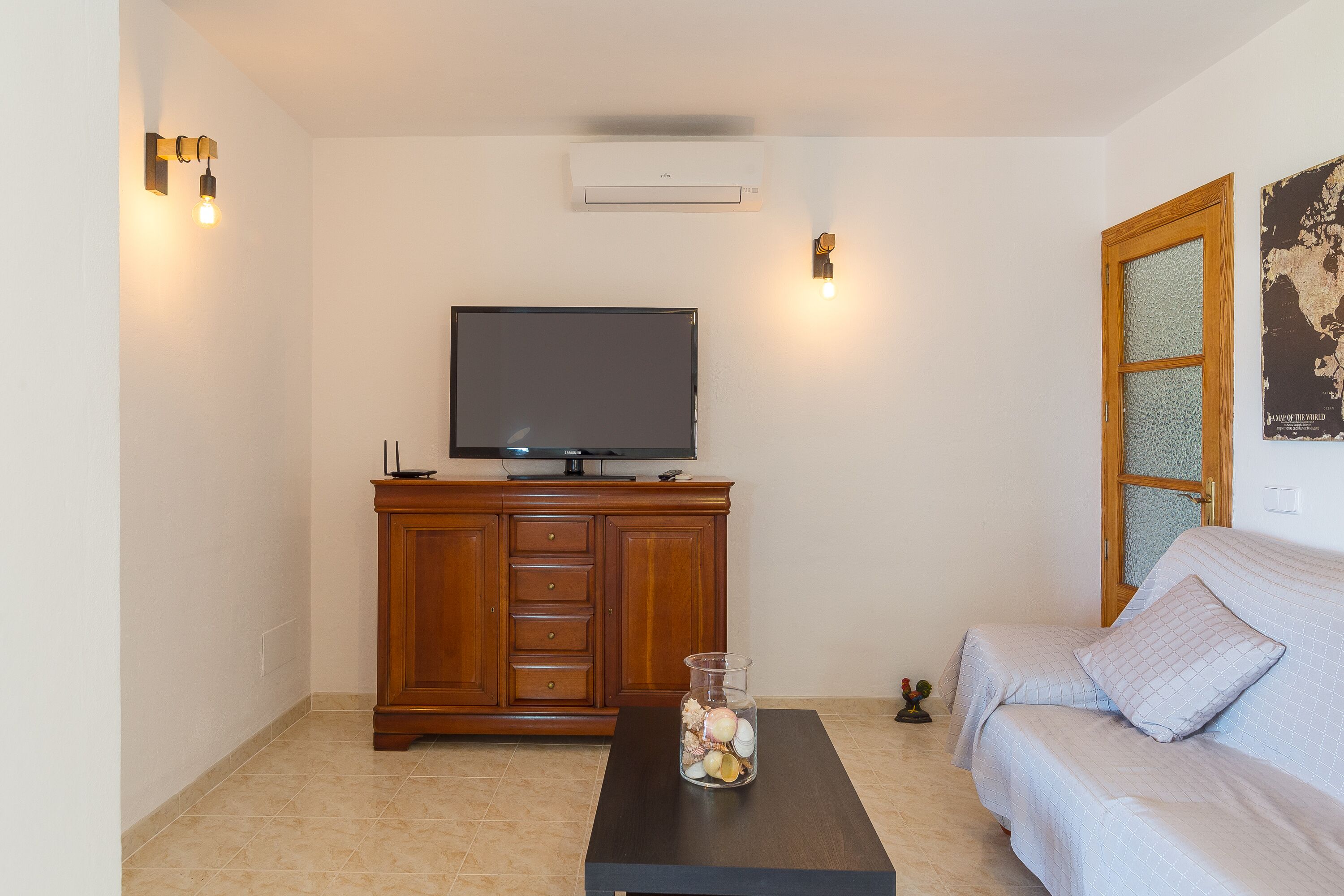 Un salon avec un téléviseur à écran plat posé sur un meuble en bois, un vase en verre contenant des objets décoratifs sur une table basse, et un canapé agrémenté d’un coussin.