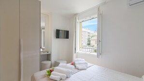 Un dormitorio con cama, mesita de noche, espejo, televisor y ventana con vistas a edificios.