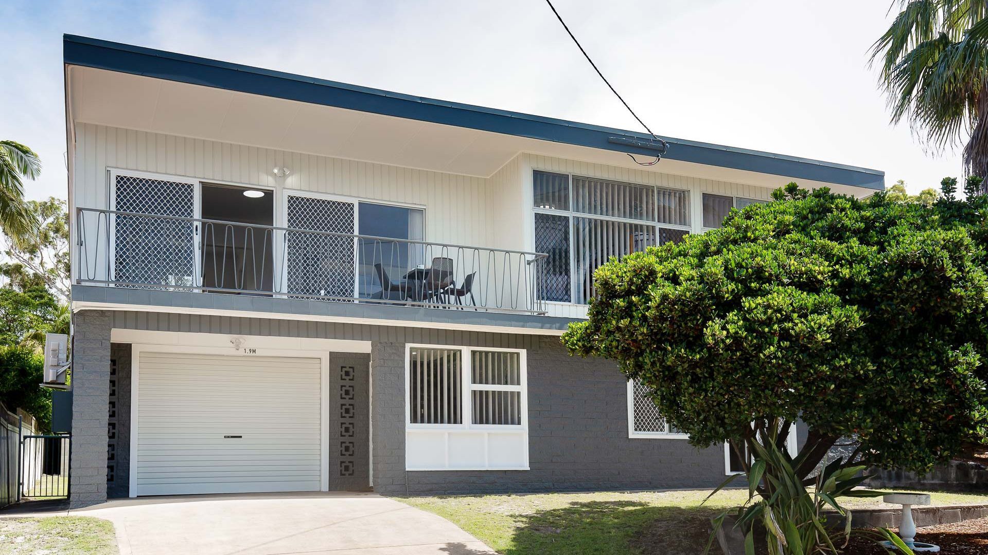 A two-story residential building with a garage, a balcony with a metal railing, and a large tree in the front yard.