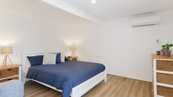 Een slaapkamer met een bed, nachtkastjes, een lamp, een wandairconditioner en een kleine plant op een bijzettafeltje.