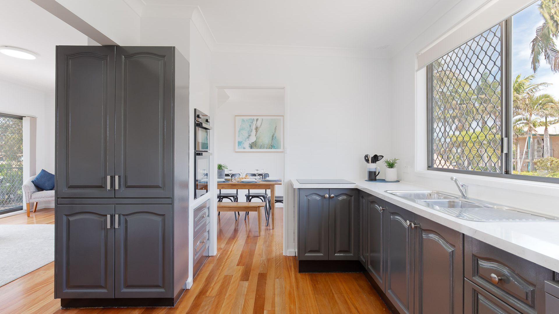 Una cocina moderna con armarios oscuros, piso de madera y un ventanal con vistas a palmeras.