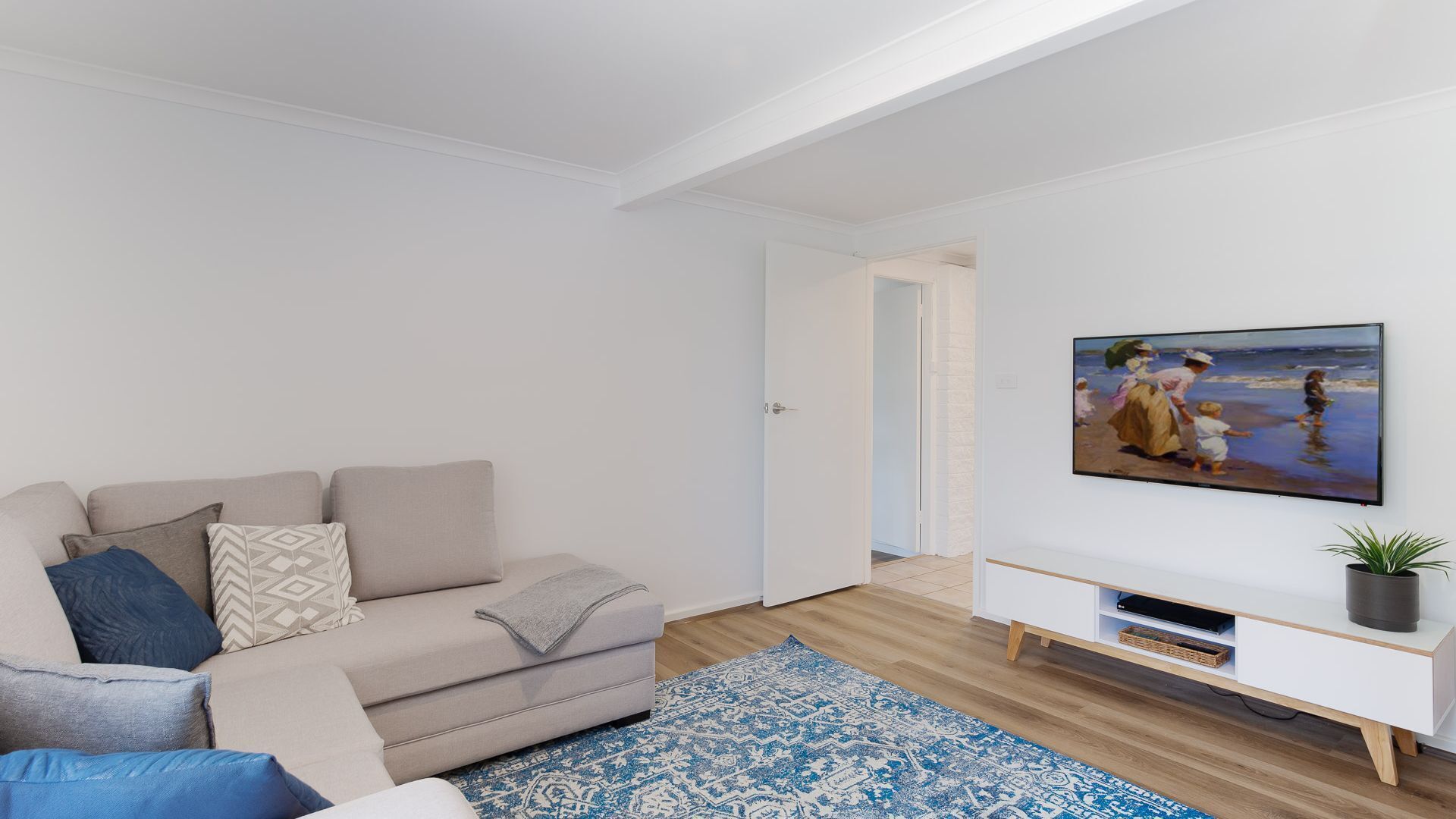 A modern living room with a beige sofa, patterned cushions, a blue rug, and a painting of a beach scene on the wall.