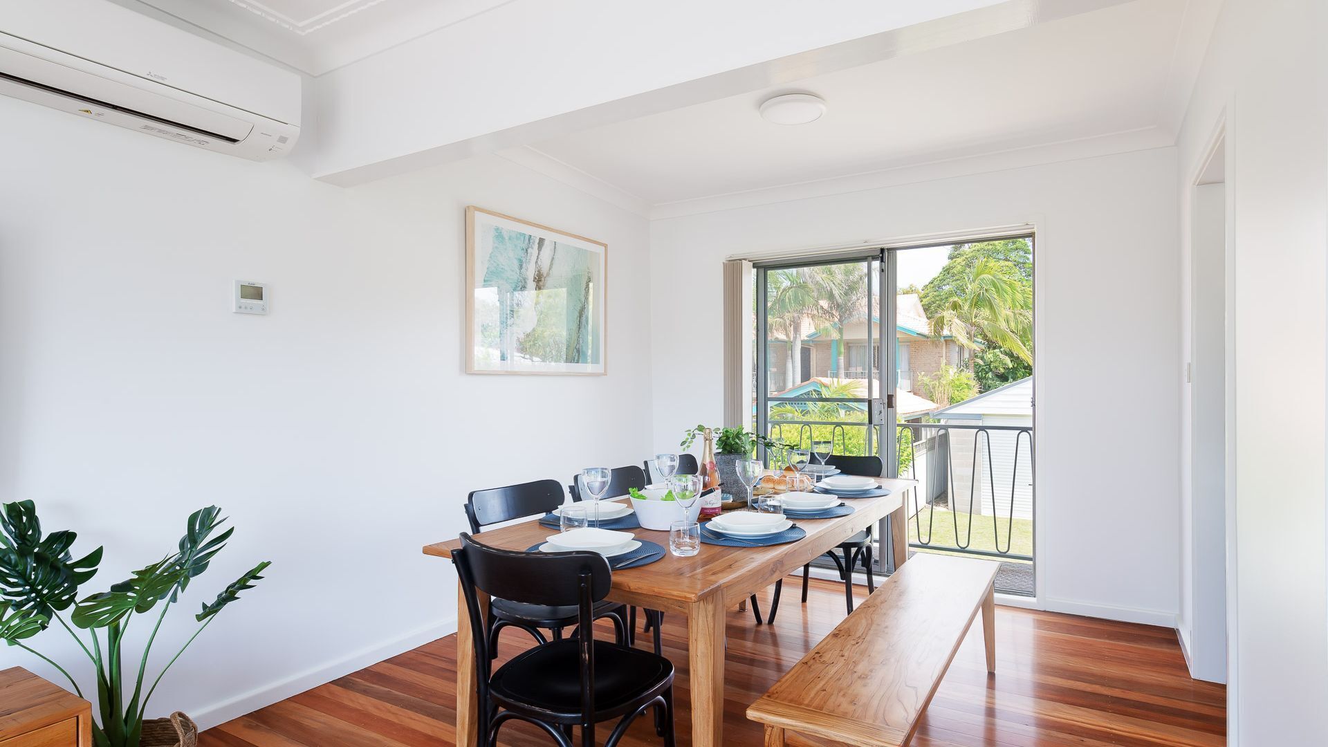 A dining area with a wooden table set for four, black chairs, and a bench. There is a window with a view of a house and greenery outside.