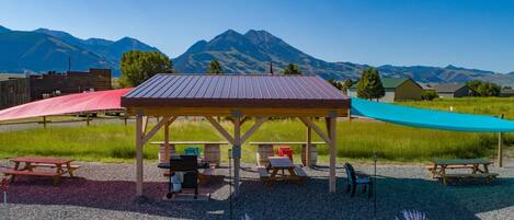 A covered outdoor seating area with picnic tables and chairs, surrounded by a gravel area with small plants, set against a backdrop of mountains.